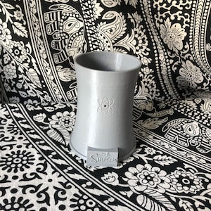 May include: A gray ceramic mug shaped like the Springfield Nuclear Power Plant from The Simpsons. The mug has a stylized atom symbol on the front and the words "The Simpsons" on the bottom.