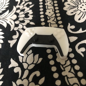 May include: A white plastic controller with black buttons and a black design. The controller is a miniature version of a video game controller.