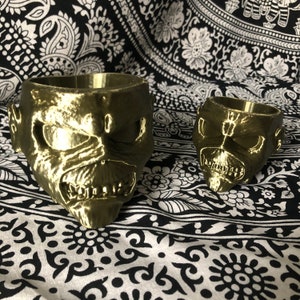 Può includere: Due vasi da fiori stampati in 3D color oro a forma di teschio con un'espressione minacciosa. Il vaso più grande è in primo piano e quello più piccolo è sullo sfondo.