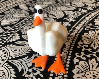 May include: A white 3D printed goose figurine with orange feet and black eyes. The goose is standing on a black and white patterned surface.