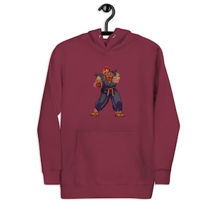 May include: Burgundy hoodie featuring a pixelated image of a character in a fighting stance. The character is wearing a blue outfit with orange hair and accents. The hoodie has a front pocket and a drawstring hood.