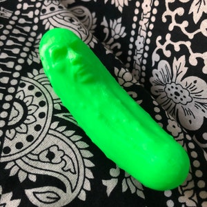 May include: A bright green pickle shaped like a human face. The pickle is lying on a black and white patterned fabric.