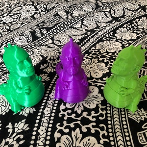 Puede incluir: Tres figuras impresas en 3D de una cabeza humana con un cuerpo de dragón. Las figuras son verdes, moradas y verdes. Las figuras están sentadas sobre una superficie con un patrón blanco y negro.