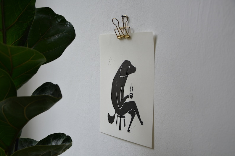 May include: Black and white illustration of a dog sitting on a stool and holding a cup of coffee. The dog is drawn in a simple, stylized manner.