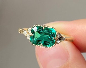 May include: A gold ring with a large emerald green gemstone surrounded by smaller diamonds. The ring is set in a delicate, vintage-inspired style.