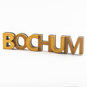 May include: A 3D-printed sign with the word "BOCHUM" in gold letters with black outlines.