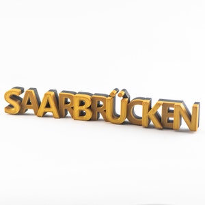 Könnte beinhalten: Ein dreidimensionales Schild mit dem Wort "SAARBRÜCKEN" in Gold und Schwarz. Die Buchstaben sind in einer fetten, serifenlosen Schriftart gehalten, mit goldener Vorderseite und schwarzen Seiten, vor weißem Hintergrund.
