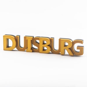 Könnte beinhalten: Ein 3D-gedrucktes Schild mit dem Wort "DUISBURG" in goldenen Buchstaben mit schwarzen Umrissen.