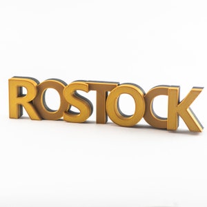 Könnte beinhalten: Das Wort "ROSTOCK" ist in 3D-Buchstaben geschrieben. Die Buchstaben sind aus goldfarbenem Material und haben einen schwarzen Umriss.