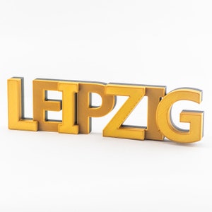 Könnte beinhalten: Ein 3D-gedrucktes Schild mit der Aufschrift "LEIPZIG" in goldenen Buchstaben mit schwarzen Umrandungen.