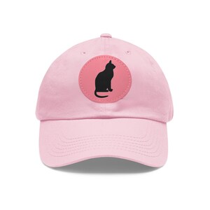 May include: A light pink baseball cap with a curved brim. A circular patch on the front features a black silhouette of a sitting cat against a pink background. The cap is made of a soft fabric.