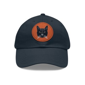 May include: Navy blue baseball cap with a round, orange patch featuring a black cat face with green eyes and white whiskers. The cap has a curved brim and a fabric strap closure.