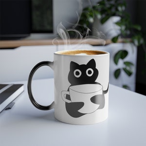 Puede incluir: Taza de cerámica blanca con asa negra e ilustración de un gato negro sosteniendo una taza de café. La taza está llena de una bebida caliente, y sale vapor de la parte superior. El gato tiene ojos grandes y redondos.