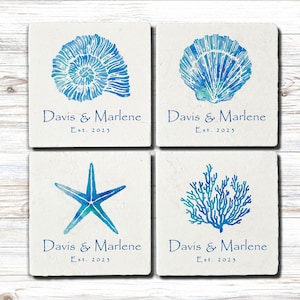 May include: Four square coasters with watercolor designs of a seashell, scallop shell, starfish, and coral in shades of blue. Each coaster has the text "Davis & Marlene Est. 2023" in a dark blue script.