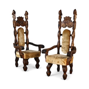 May include: Two ornate, dark wood chairs with high backs and armrests. The chairs have carved details, including figures at the top of the backrests. The seats and backrests are upholstered in a light gold fabric. The chairs are on a white background.