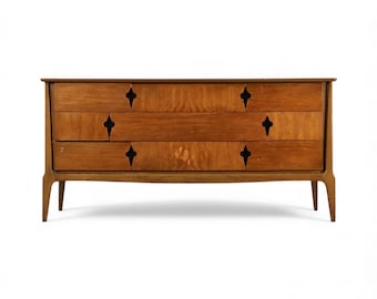 May include: A mid-century modern wooden dresser with three drawers. The dresser is made of light brown wood with a natural grain pattern. Each drawer has a decorative diamond-shaped cutout. The dresser has four tapered legs.