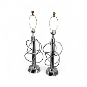 May include: Pair of chrome table lamps with a unique, looping design. Each lamp features a cylindrical base, a central pole, and a decorative metalwork element. The lamps have a silver finish and are topped with a light bulb fixture.