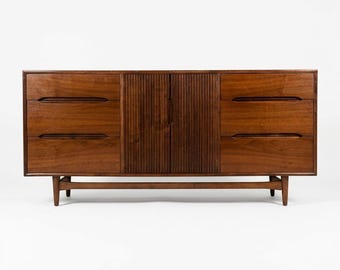 May include: A mid-century modern dresser with a warm brown finish. The dresser features six drawers and two doors with vertical grooved detailing. It stands on tapered legs and has a long, horizontal design.