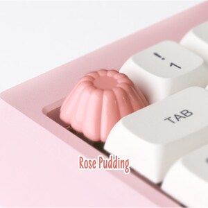 May include: A pink keyboard with a rose pudding keycap. The keycap is shaped like a small cake with a pink frosting.