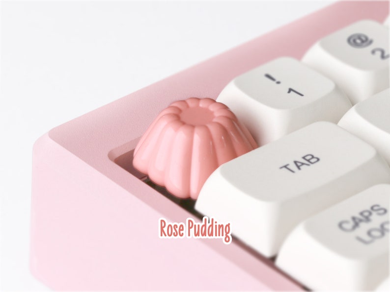 May include: A pink keyboard with a rose pudding keycap. The keycap is shaped like a small cake with a pink frosting.