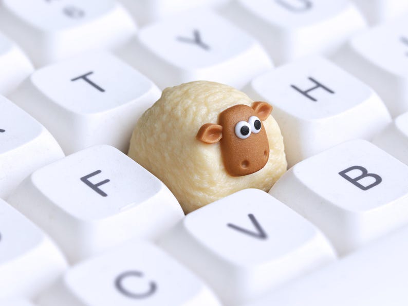 May include: A close-up image of a sheep figurine resting on a white computer keyboard. The sheep is a light beige colour with a brown face and ears, and large, googly eyes. The keyboard keys are labelled with black letters.