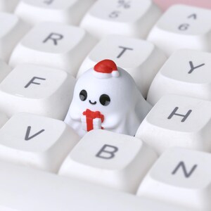 K&ouml;nnte beinhalten: Eine wei&szlig;e Tastatur mit wei&szlig;en Tastenkappen. Eine individuelle Tastenkappe in Form eines Geistes mit Weihnachtsm&uuml;tze und rotem Geschenk befindet sich auf der Tastatur. Die Tastenkappe hat schwarze Augen und einen l&auml;chelnden Mund.