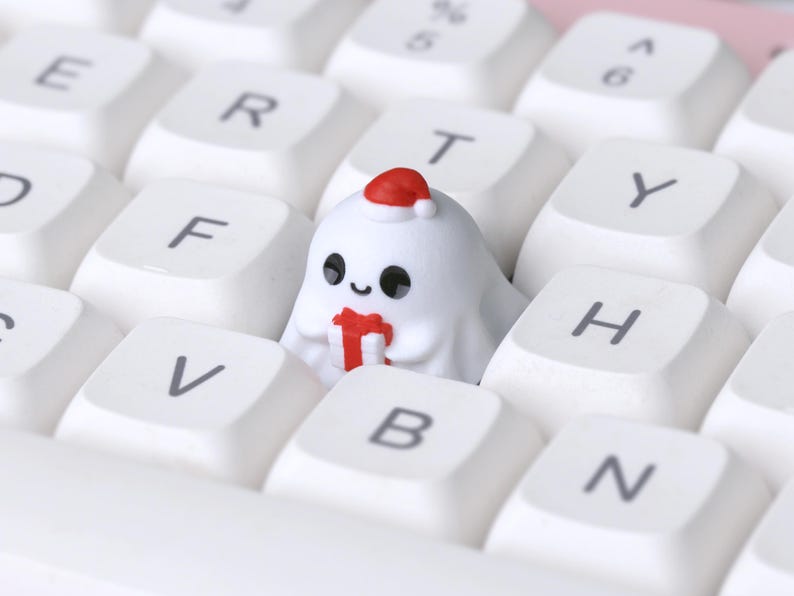 K&ouml;nnte beinhalten: Eine wei&szlig;e Tastatur mit wei&szlig;en Tastenkappen. Eine individuelle Tastenkappe in Form eines Geistes mit Weihnachtsm&uuml;tze und rotem Geschenk befindet sich auf der Tastatur. Die Tastenkappe hat schwarze Augen und einen l&auml;chelnden Mund.