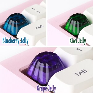 May include: Three jelly keycaps in different colors: blue, green, and purple. The keycaps are shaped like a jelly mold and are sitting on a white keyboard.