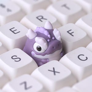 May include: A purple dragon-shaped artisan keycap featuring large black eyes, white horns, and a textured crest, positioned between the white keys of a computer keyboard with visible letters including E, W, S, F, C, and X.