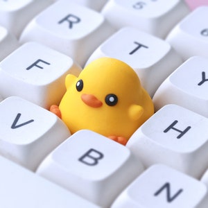 Pode incluir: Um brinquedo de pintinho amarelo sentado em um teclado branco com teclas rotuladas como 'V', 'B', 'N', 'H', 'Y', 'T', 'R' e 'F'.