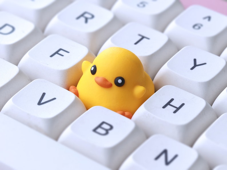Pode incluir: Um brinquedo de pintinho amarelo sentado em um teclado branco com teclas rotuladas como 'V', 'B', 'N', 'H', 'Y', 'T', 'R' e 'F'.