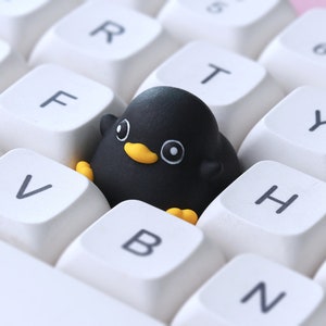 Pode incluir: Uma borracha preta em forma de pinguim est&aacute; sobre um teclado branco com teclas rotuladas como 'V', 'B', 'N', 'H', 'Y', 'T', 'R', 'F' e 'D'.