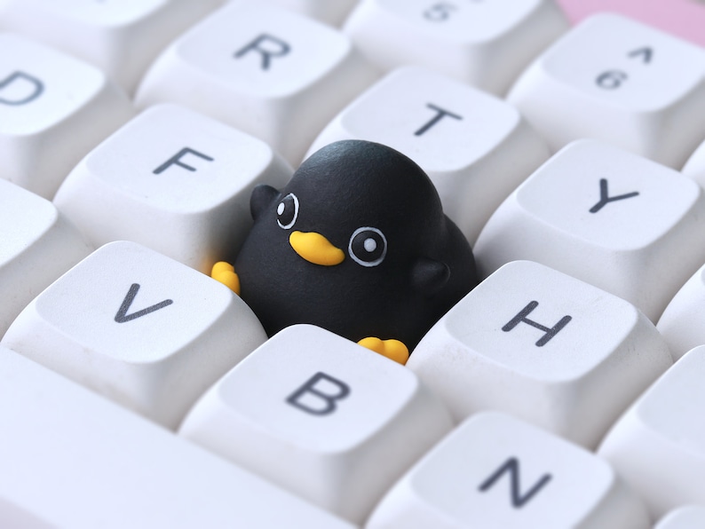 Pode incluir: Uma borracha preta em forma de pinguim est&aacute; sobre um teclado branco com teclas rotuladas como 'V', 'B', 'N', 'H', 'Y', 'T', 'R', 'F' e 'D'.