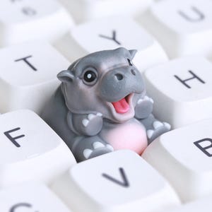 Puede incluir: Una peque&ntilde;a figura de hipop&oacute;tamo gris sentada en un teclado de computadora blanco. El hipop&oacute;tamo est&aacute; sonriendo y tiene la boca abierta.