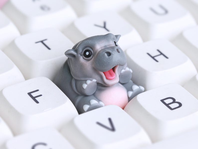 Puede incluir: Una peque&ntilde;a figura de hipop&oacute;tamo gris sentada en un teclado de computadora blanco. El hipop&oacute;tamo est&aacute; sonriendo y tiene la boca abierta.