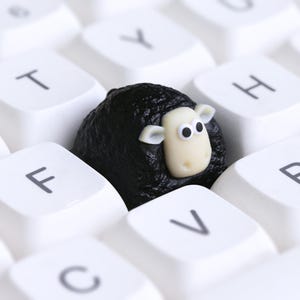 May include: A black sheep ornament with a white face and ears sits amongst white keyboard keys. The sheep is surrounded by white keys with black letters. The sheep's face has two black eyes.