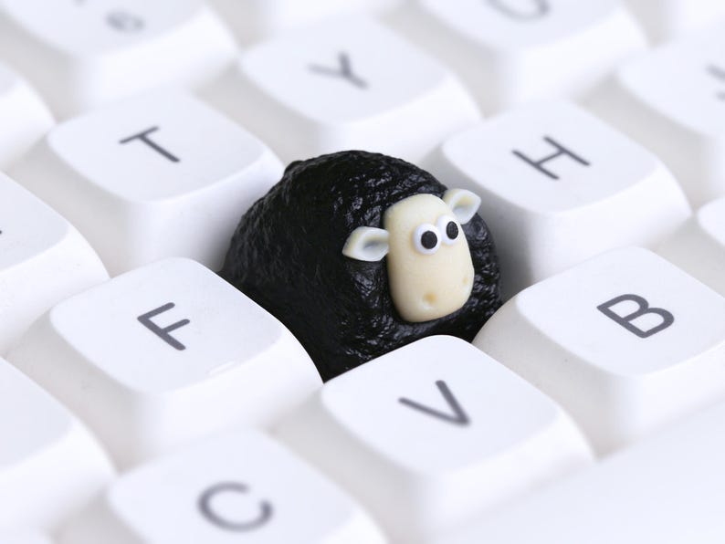 May include: A black sheep ornament with a white face and ears sits amongst white keyboard keys. The sheep is surrounded by white keys with black letters. The sheep's face has two black eyes.