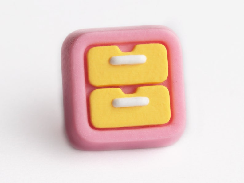 May include: A pink square-shaped drawer pull with two yellow drawers and white handles. The drawer pull has a rounded edge and is set against a white background. The design is simple and colorful.