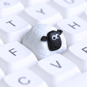 May include: A white keyboard featuring a novelty sheep-shaped keycap. The sheep keycap has a white body and a black face with large eyes. The keyboard keys are white with black lettering.