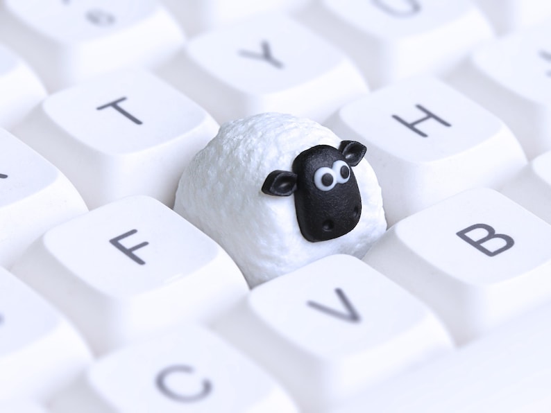 May include: A white keyboard featuring a novelty sheep-shaped keycap. The sheep keycap has a white body and a black face with large eyes. The keyboard keys are white with black lettering.