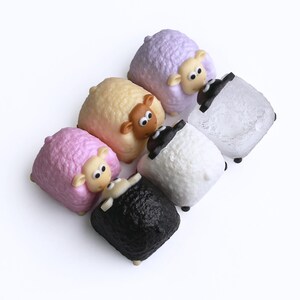 May include: Six sheep figurines in pastel shades of pink, yellow, and lavender, alongside white and black sheep. Each has a cartoon face with large eyes and small ears. The figurines are arranged on a white surface.