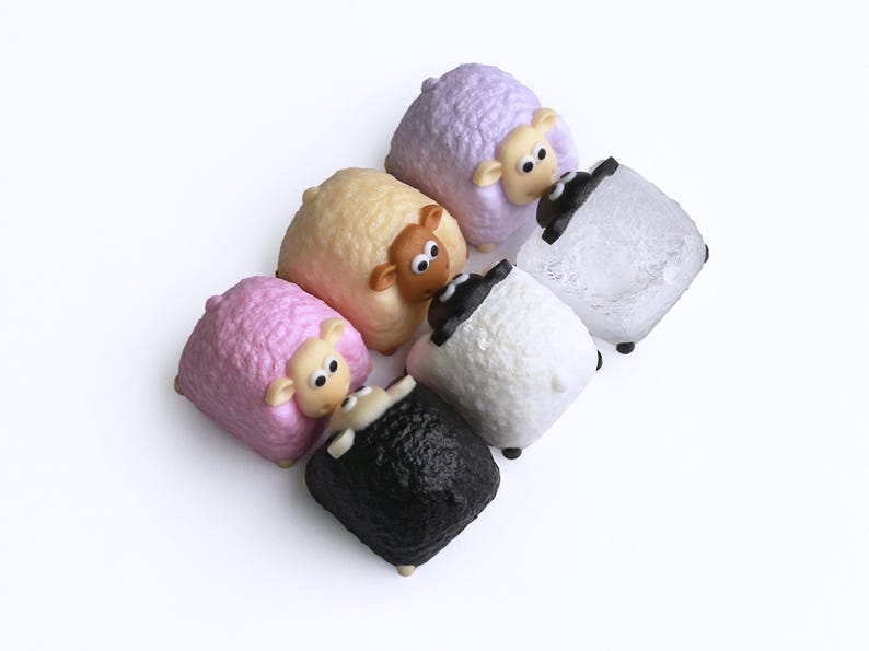 May include: Six sheep figurines in pastel shades of pink, yellow, and lavender, alongside white and black sheep. Each has a cartoon face with large eyes and small ears. The figurines are arranged on a white surface.
