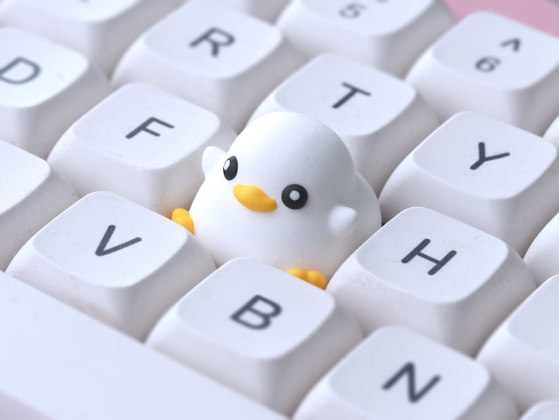 Pode incluir: Um teclado branco com uma pequena figura de pintinho branco sentada nas teclas F, V e T.