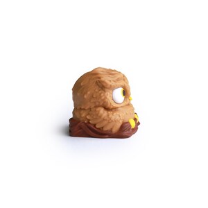 Puede incluir: Figura de b&uacute;ho marr&oacute;n con superficie texturizada, ojo grande blanco y amarillo, y detalles amarillos en las patas. El b&uacute;ho est&aacute; posado sobre una base marr&oacute;n, sobre fondo blanco.