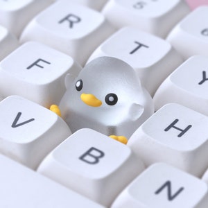 Pode incluir: Um teclado branco com uma figura de pinguim de pl&aacute;stico transparente sentada nas teclas F, V e B.