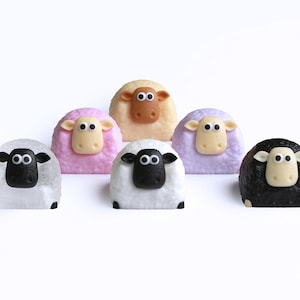 May include: Six sheep-shaped objects in a variety of colours, including white, pink, tan, and black. Each sheep has a contrasting face and ears, with black eyes. The objects are set against a white background.