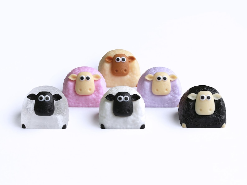 May include: Six sheep-shaped objects in a variety of colours, including white, pink, tan, and black. Each sheep has a contrasting face and ears, with black eyes. The objects are set against a white background.