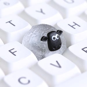 May include: A novelty sheep-shaped keycap sits amongst white keyboard keys. The sheep is grey with a black face and white eyes. The keyboard keys have black lettering. A fun, quirky design.