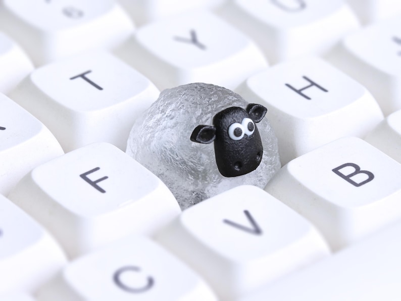 May include: A novelty sheep-shaped keycap sits amongst white keyboard keys. The sheep is grey with a black face and white eyes. The keyboard keys have black lettering. A fun, quirky design.