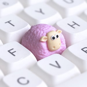 May include: A close-up of a white computer keyboard with a pink sheep-shaped object in the center. The sheep has a light beige face and black eyes. The keyboard keys are labelled with letters.